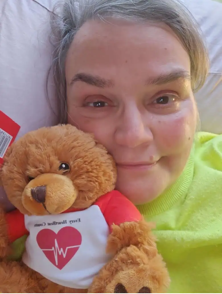 A woman smiling and holding a brown teddy bear with a heart logo shirt.