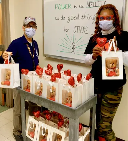 Two masked individuals holding gift bags with red bows beside a cart full of similar bags.